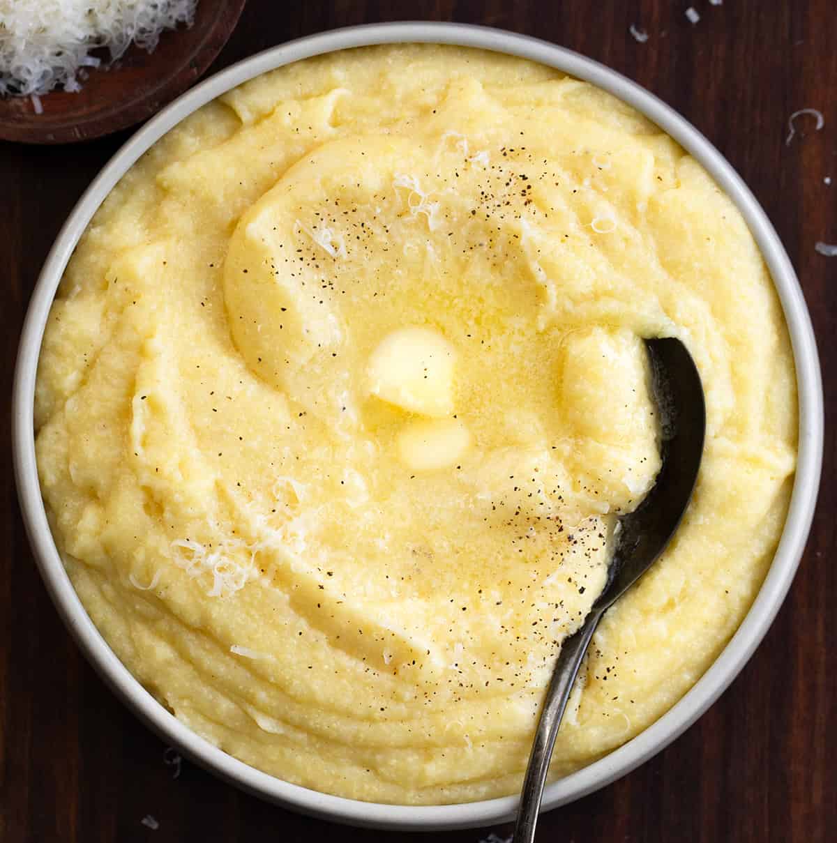 Creamy polenta is comfort food made simple. Slowly cooked fine cornmeal turns buttery and smooth, creating the perfect base for hearty meals or cozy weeknight dinners! Bowl of Creamy Polenta on a wooden table with a spoon in it.