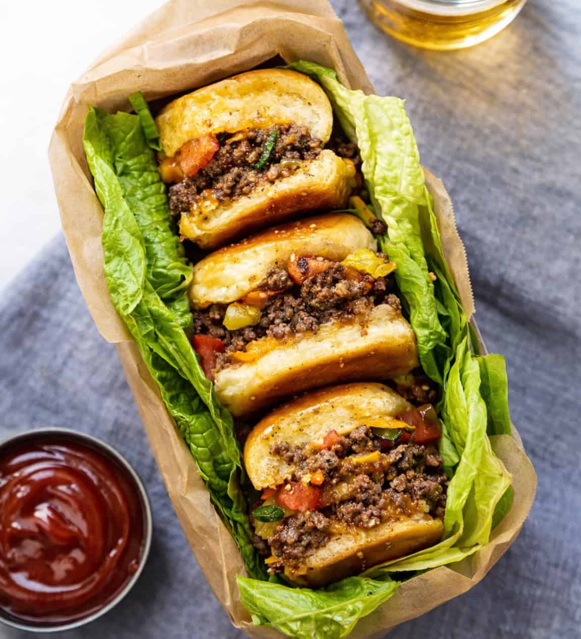 Sliders are one of the easiest ways to feed a crowd. They’re fun, customizable, and perfect for parties, game days, holidays, and family dinners! Cheeseburgers sliders in a pan with lettuce on a table with ketchup and beer.