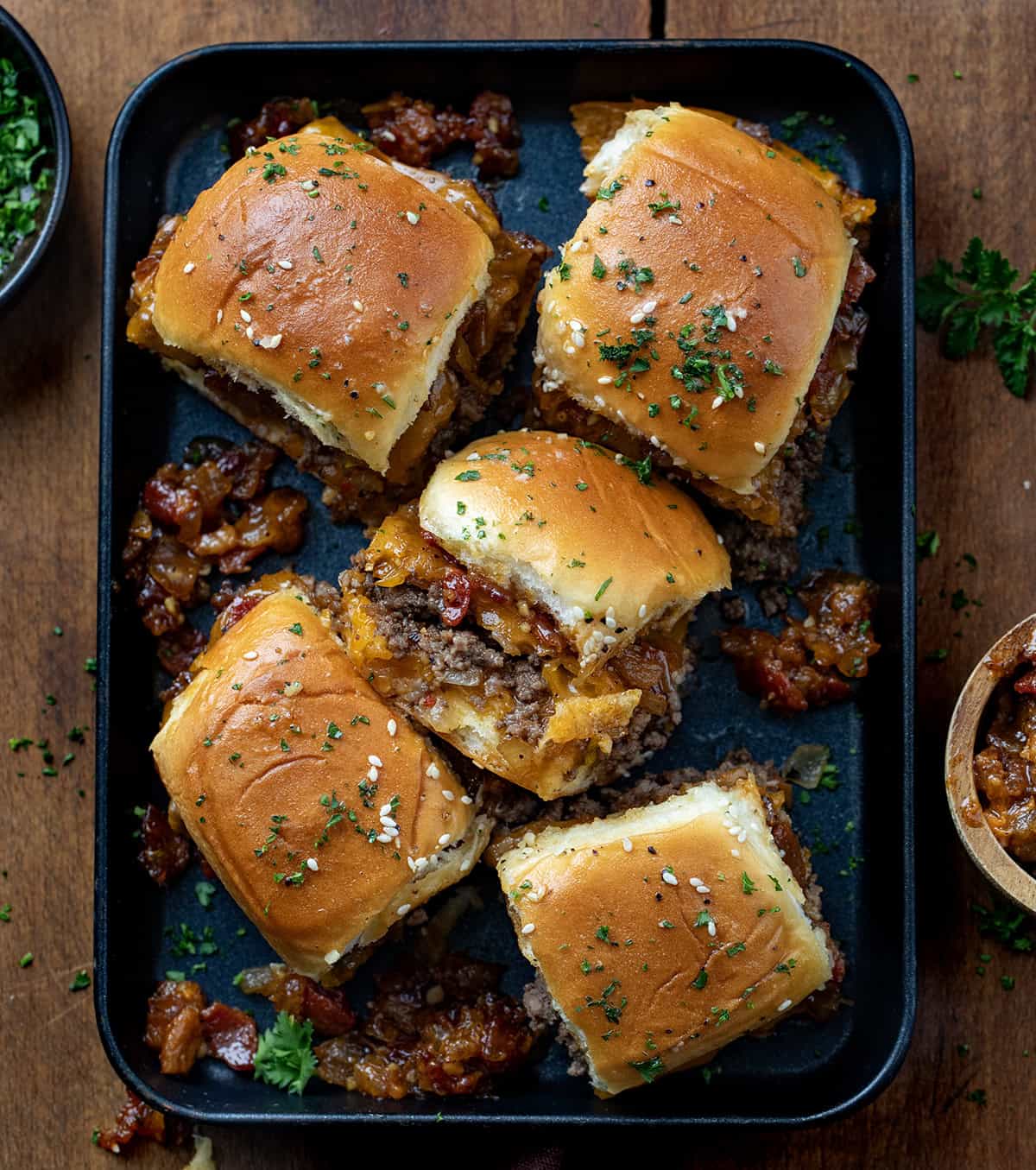 These Bacon Jam Cheeseburger Sliders are soft, sweet Hawaiian rolls layered with juicy, seasoned ground beef, melty cheddar, and smoky homemade bacon jam. Bacon Jam Cheeseburger Sliders in a black pan on a wooden table from overhead.