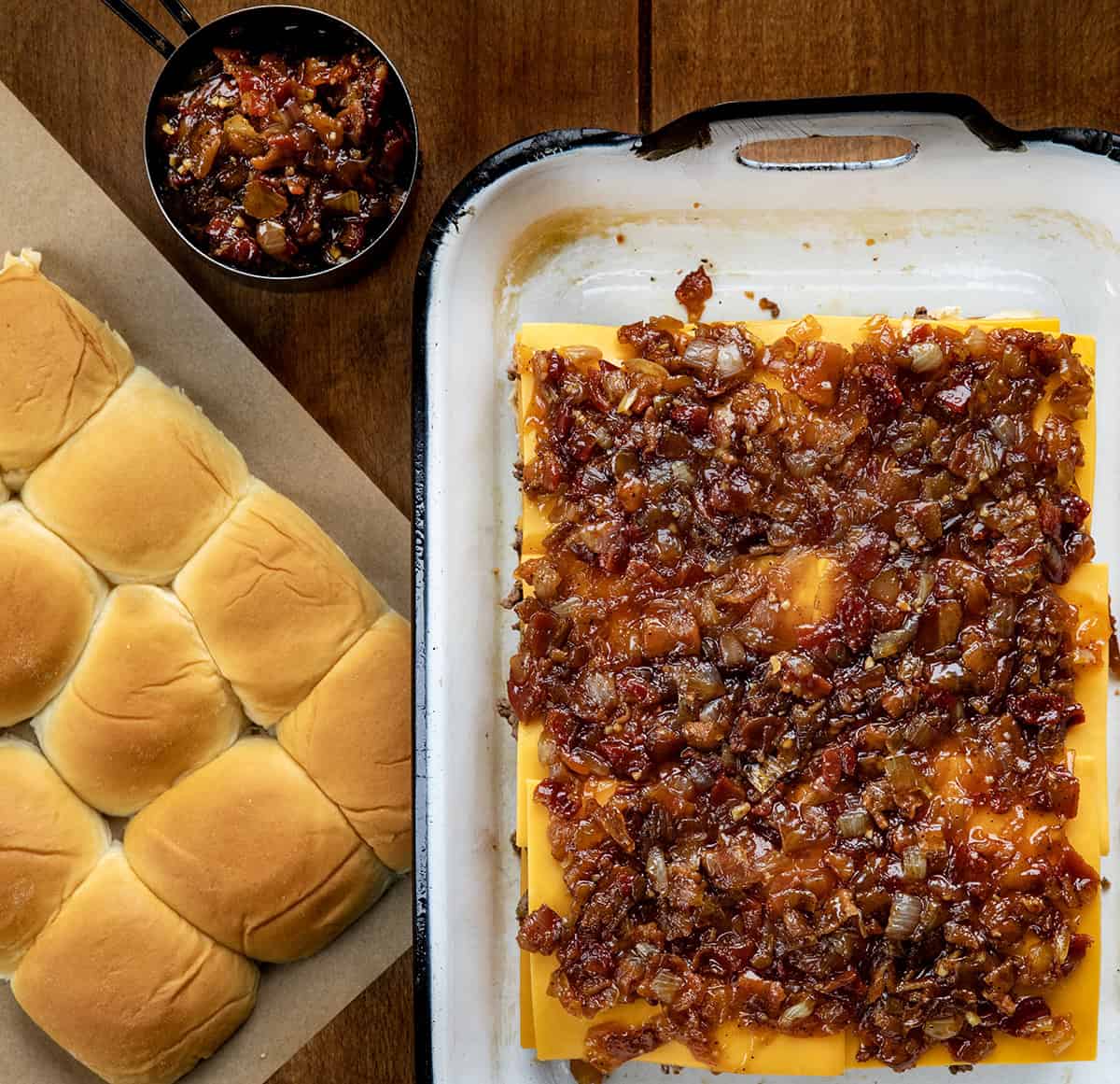 These Bacon Jam Cheeseburger Sliders are soft, sweet Hawaiian rolls layered with juicy, seasoned ground beef, melty cheddar, and smoky homemade bacon jam. Pan of almost finished Bacon Jam Cheeseburger Sliders with top bun and bacon jam off to the side.