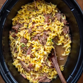 Slow Cooker filled with Beef and Noodles with a wooden spoon on a wooden table.