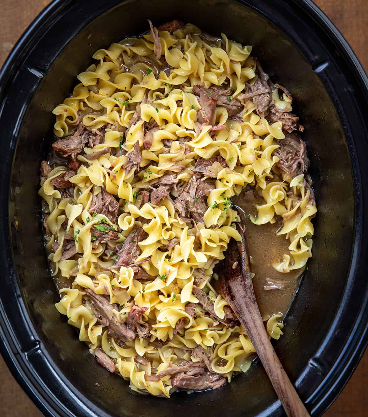 This Slow Cooker Beef and Noodles is a cozy, family-friendly dinner with tender, fall-apart beef, rich savory gravy, and soft egg noodles. Easy to make and perfect for busy weeknights or a comforting weekend meal. Slow Cooker filled with Beef and Noodles with a wooden spoon on a wooden table.