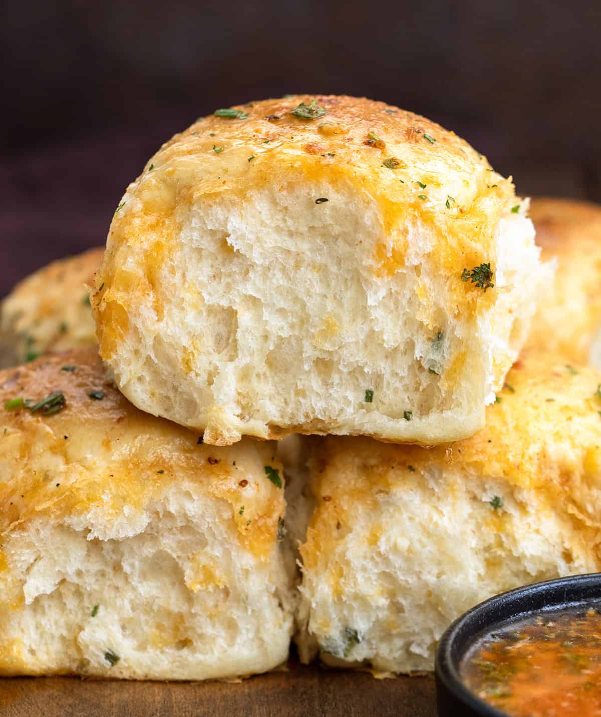 These Cheesy Cowboy Butter Dinner Rolls are the best of both worlds: a mash-up of my Cheesy Garlic Dinner Rolls and my popular Cowboy Butter Dinner Rolls (without cheese), both of which are amazing on their own! Stacked Cheesy Cowboy Butter Dinner Rolls on a wooden table next to cowboy butter.