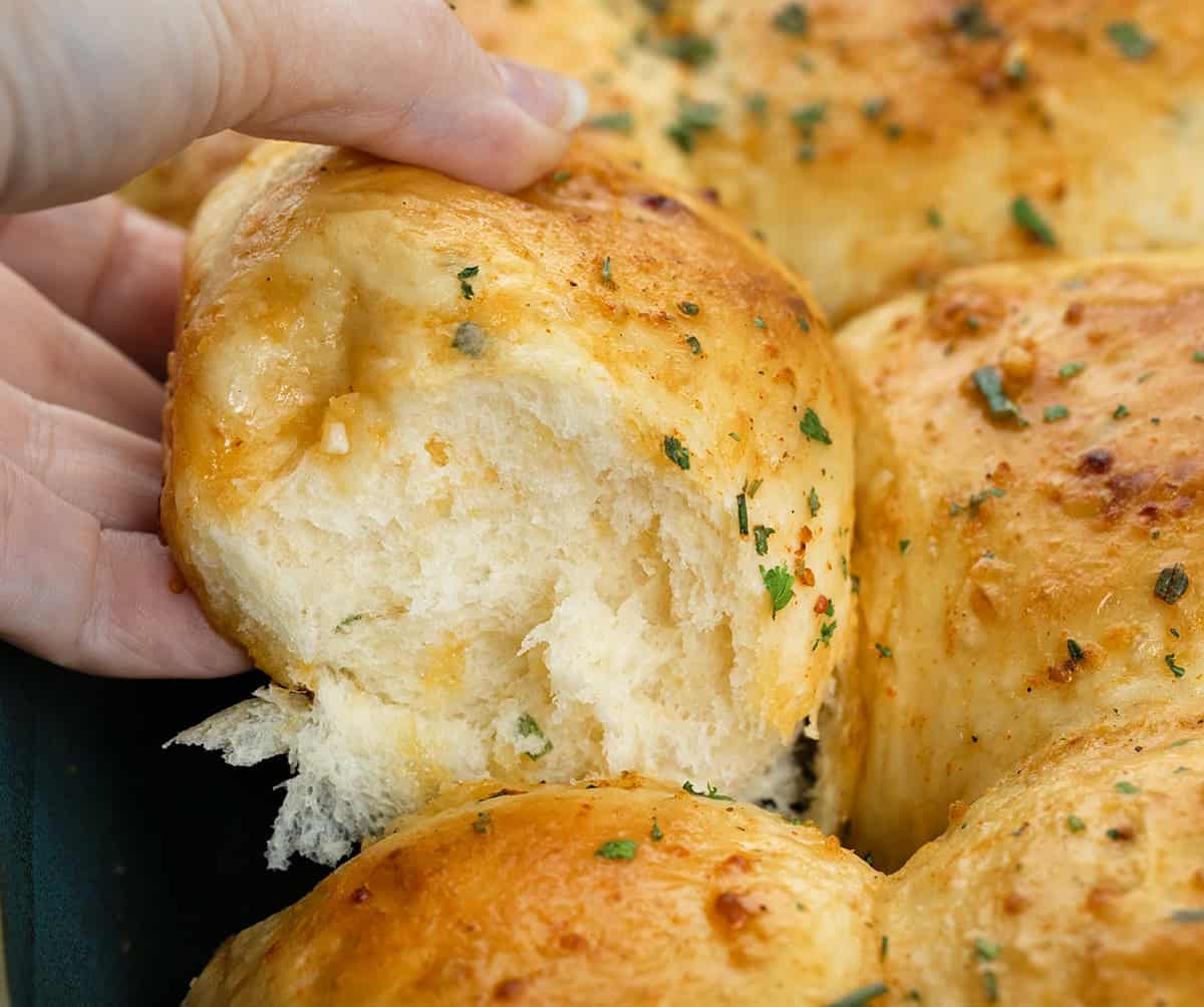 These Cheesy Cowboy Butter Dinner Rolls are the best of both worlds: a mash-up of my Cheesy Garlic Dinner Rolls and my popular Cowboy Butter Dinner Rolls (without cheese), both of which are amazing on their own! Hand picking up a Cheesy Cowboy Butter Dinner Roll from the pan.