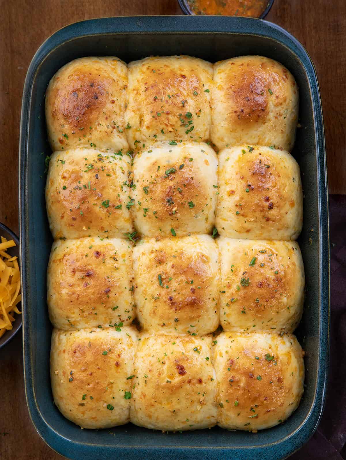 These Cheesy Cowboy Butter Dinner Rolls are the best of both worlds: a mash-up of my Cheesy Garlic Dinner Rolls and my popular Cowboy Butter Dinner Rolls (without cheese), both of which are amazing on their own! Pan of Cheesy Cowboy Butter Dinner Rolls on a wooden table from overhead.