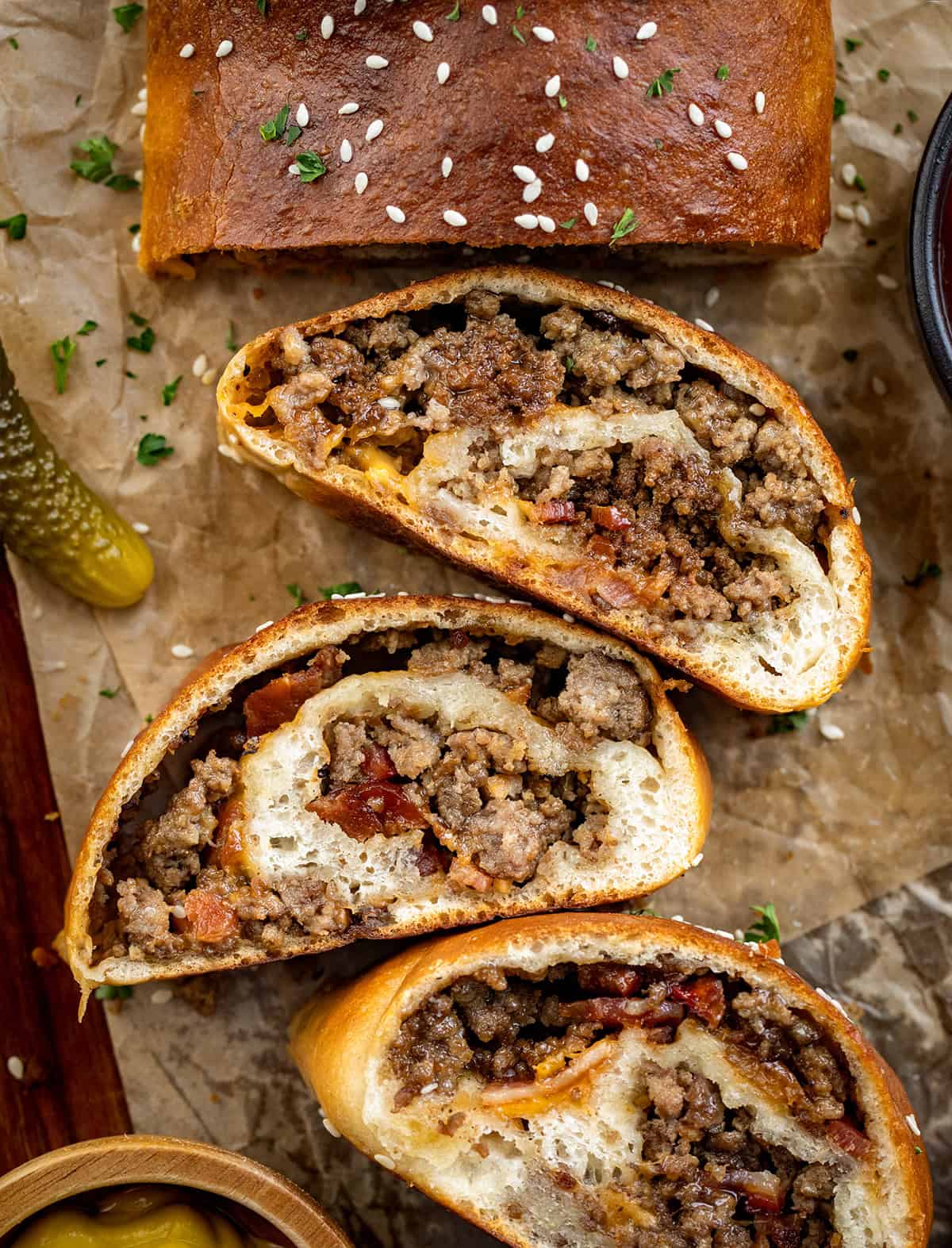 Bacon Cheeseburger Garbage Bread is packed with seasoned beef, crispy bacon, and melted cheddar, all rolled up in golden dough. Slice and serve warm with your favorite dipping sauces for a fun family dinner or game-day snack! Garbage Bread with some slices cut and laying falt with ketchup and a pickle.