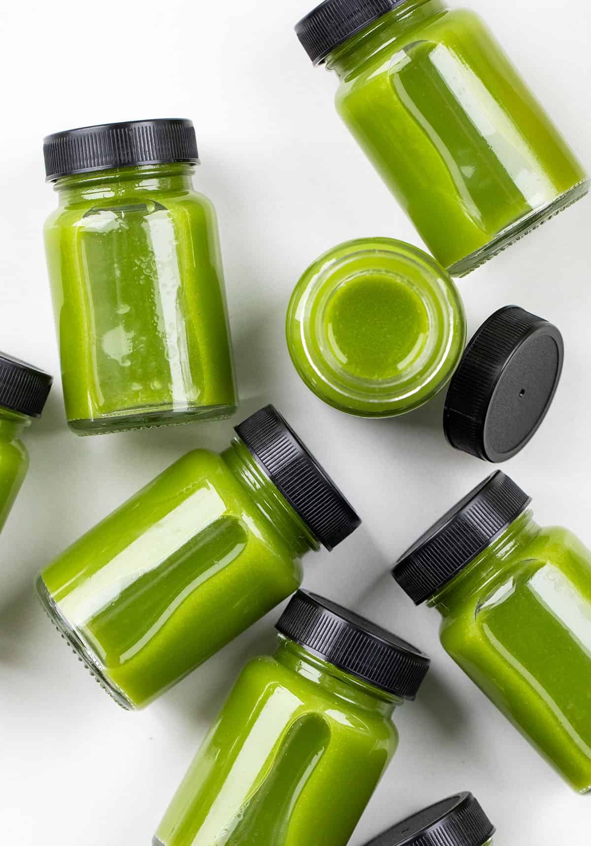 Keep your wellness routine fresh and bright with this Greens Immunity Booster! Made with crisp apples, leafy greens, zesty lemon, and a little spicy ginger, these shots are a simple, refreshing way to add a little natural energy to your day! Greens Immunity Booster on a white table with some jars on their side and one jar open.