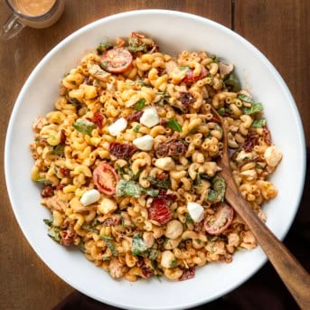White bowl filled with Marry Me Macaroni Pasta Salad and a wooden spoon in it on a wooden table.