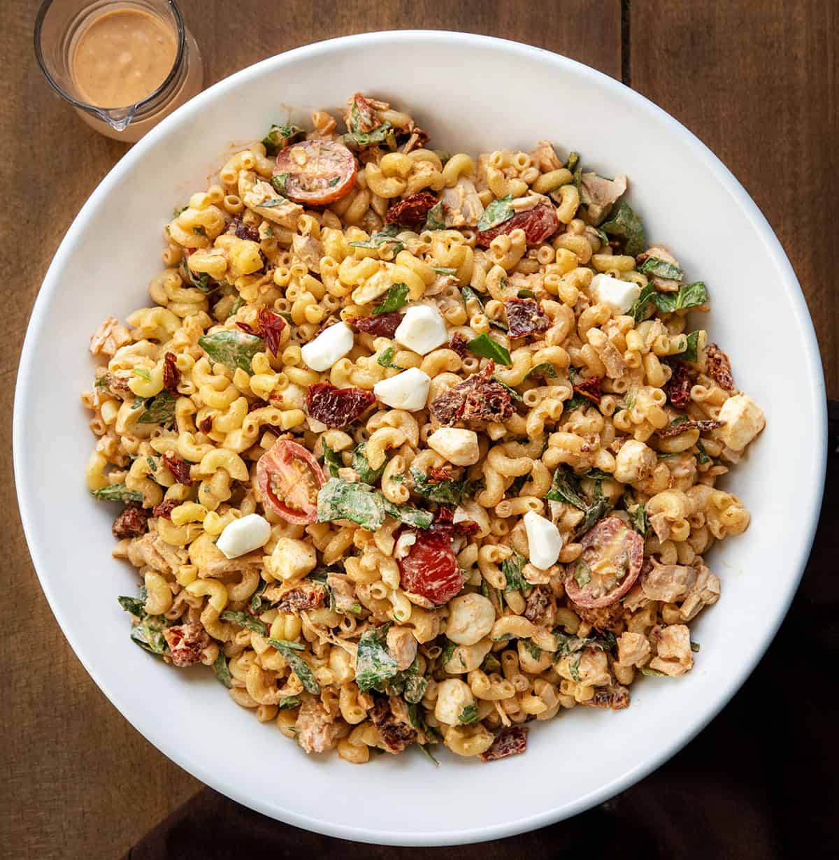 This Marry Me Pasta Salad is creamy, flavorful, and packed with tender rotisserie chicken, sun-dried tomatoes, fresh spinach, and cheesy goodness! White bowl of Marry Me Macaroni Pasta Salad on a wooden table from overhead.
