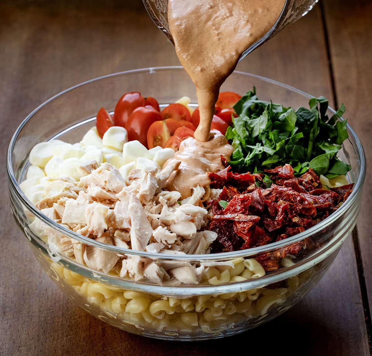 This Marry Me Pasta Salad is creamy, flavorful, and packed with tender rotisserie chicken, sun-dried tomatoes, fresh spinach, and cheesy goodness! Pouring dressing over a bowl filled with sun dreid tomatoes, noodles, chicken, mozzarella, tomatoes, and spinach.