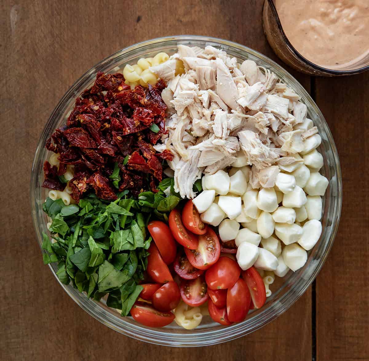 This Marry Me Pasta Salad is creamy, flavorful, and packed with tender rotisserie chicken, sun-dried tomatoes, fresh spinach, and cheesy goodness! Bowl of macaroni noodles, tomatoes, mozzarella pearls, spinach, sun-dried tomatoes, and chicken on a wooden table from overhead.