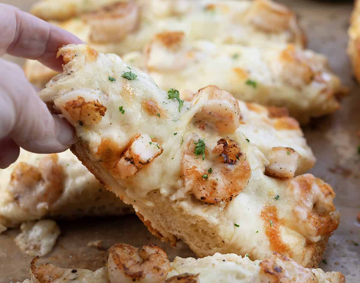 Golden, garlicky bread topped with juicy shrimp, creamy Alfredo sauce, and a gooey blend of mozzarella and white cheddar. Hand holding a piece of Shrimp Alfredo Garlic Bread.