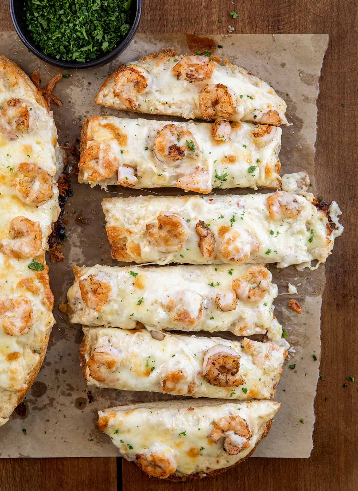 Golden, garlicky bread topped with juicy shrimp, creamy Alfredo sauce, and a gooey blend of mozzarella and white cheddar. Sliced Shrimp Alfredo Garlic Bread on a wooden table from overhead.