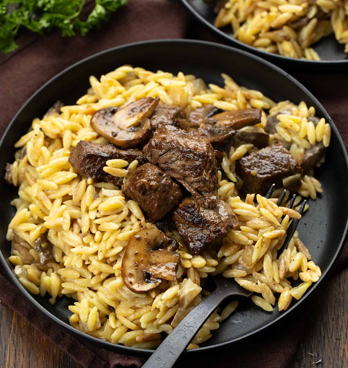 Black plate with Steak Bites with Creamy Mushroom Orzo on it and a black fork resting on the plate.