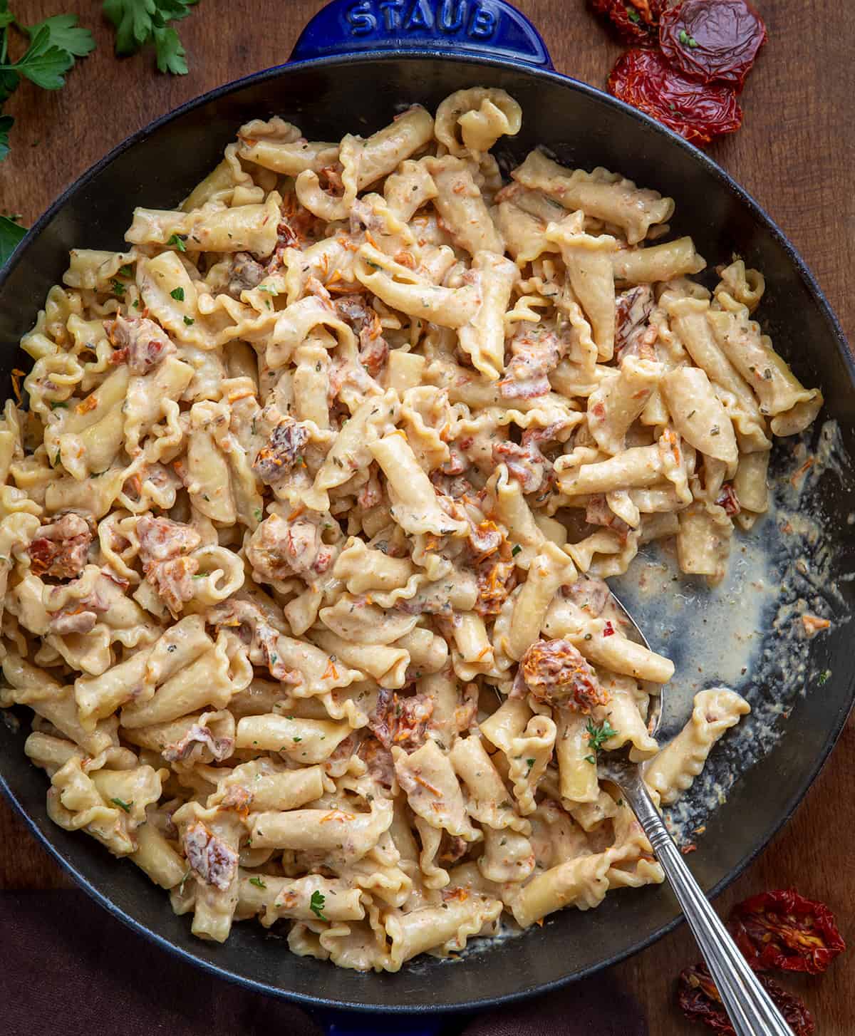 Creamy Sun-Dried Tomato Pasta is a quick and flavorful pasta with sun-dried tomatoes, garlic, Parmesan, and Tuscan seasoning. Ready in under 30 minutes, this comforting dish is perfect for weeknight dinners. Skillet of Sun-Dried Tomato Pasta with a spoon in it on a wooden table from overhead.