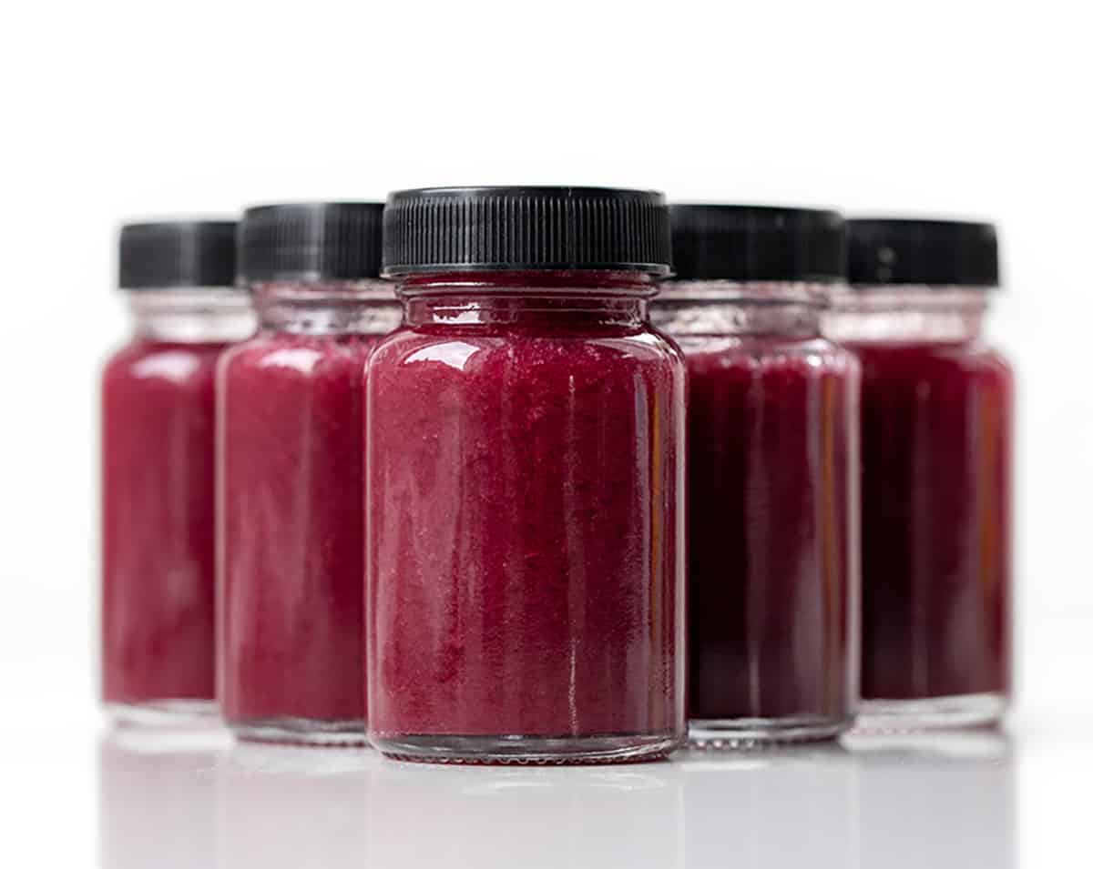 Superfood Blueberry Lemon Shots are a quick, refreshing wellness drink packed with antioxidants. Made with fresh blueberries, zesty lemon, and a hint of ginger, these healthy shots are naturally sweetened with honey and blended with coconut water. Shot glasses with black lids on filled with Superfood Blueberry Lemon Shot on a white table lined up.