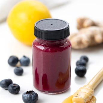 Small bottle filed with Superfood Blueberry Lemon Shot and surrounded by blueberries, lemon, ginger, and honey.
