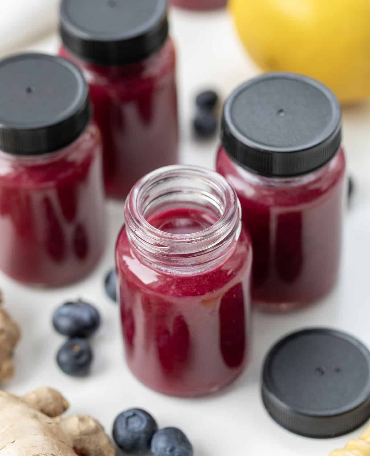 Superfood Blueberry Lemon Shots are a quick, refreshing wellness drink packed with antioxidants. Made with fresh blueberries, zesty lemon, and a hint of ginger, these healthy shots are naturally sweetened with honey and blended with coconut water. Bottles of Superfood Blueberry Lemon Shot with one bottle open on a white table.