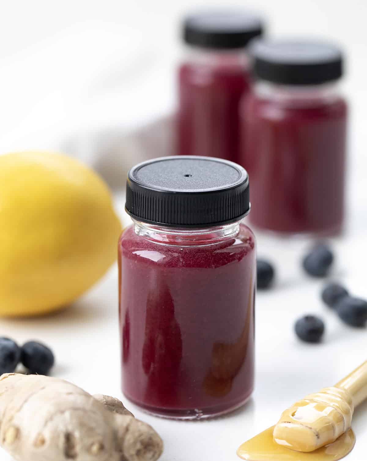 Superfood Blueberry Lemon Shots are a quick, refreshing wellness drink packed with antioxidants. Made with fresh blueberries, zesty lemon, and a hint of ginger, these healthy shots are naturally sweetened with honey and blended with coconut water. Bottles of Superfood Blueberry Lemon Shot on a white table with lemon, blueberries, and ginger.