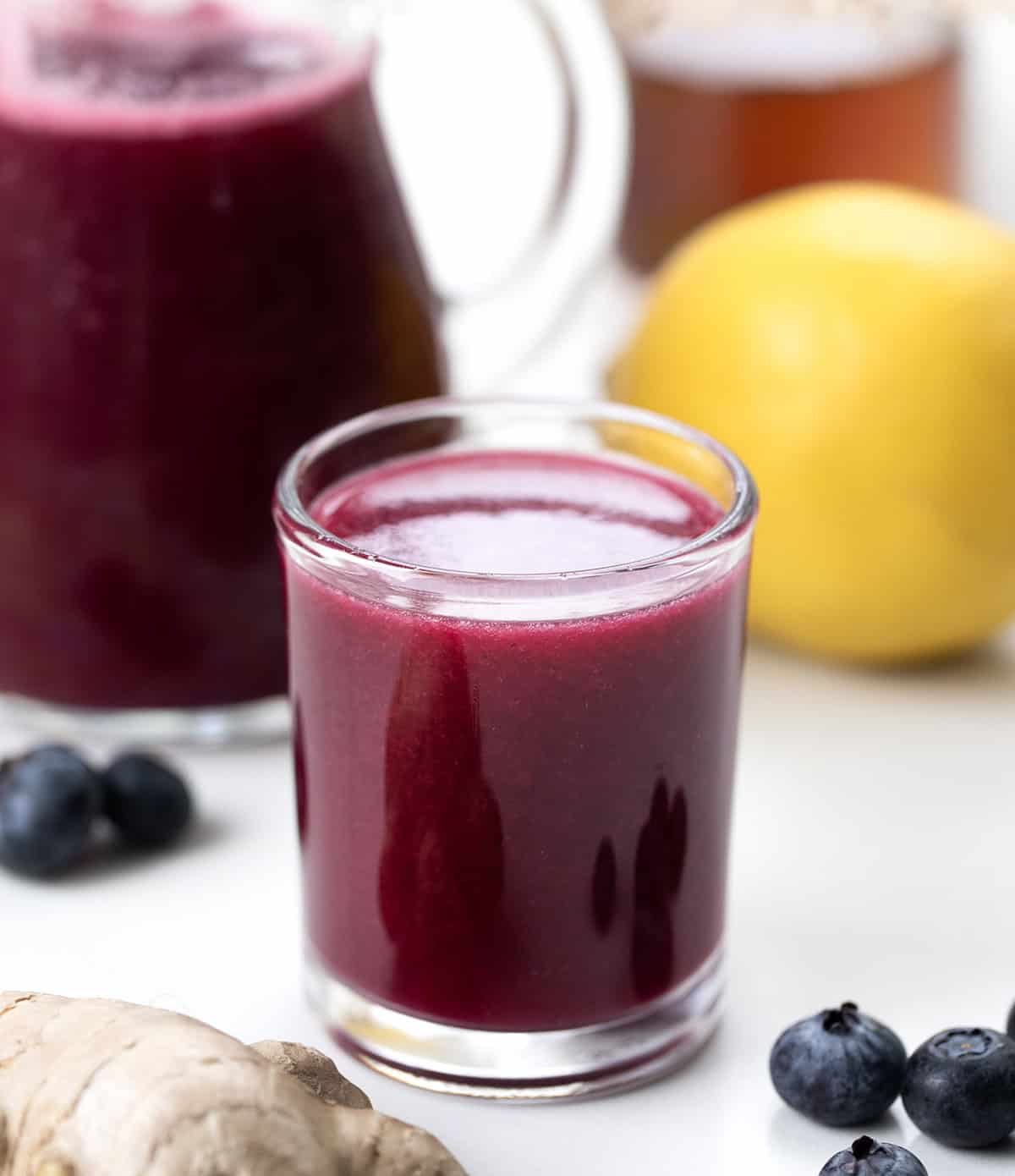 Superfood Blueberry Lemon Shots are a quick, refreshing wellness drink packed with antioxidants. Made with fresh blueberries, zesty lemon, and a hint of ginger, these healthy shots are naturally sweetened with honey and blended with coconut water. Shot glass of Superfood Blueberry Lemon Shot on a white table.