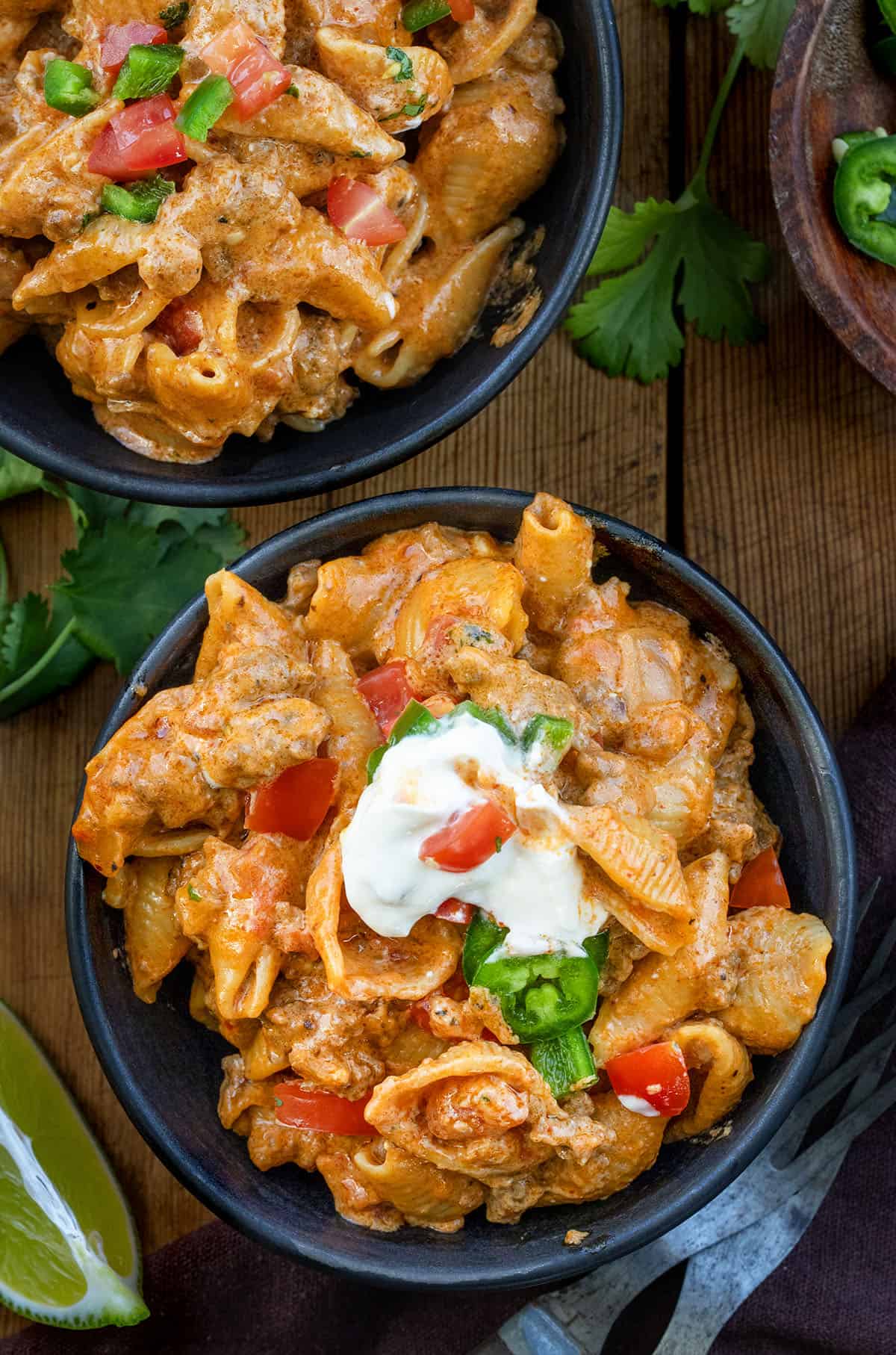 Get dinner on the table fast with this creamy, cheesy one-pot taco pasta! Packed with seasoned ground beef, tender shell pasta, and a rich, savory sauce, it’s a comforting taco-inspired meal the whole family will love on busy weeknights! Small black bowls of Taco Pasta on a wooden table from overhead with fresh lime wedges and cilantro.