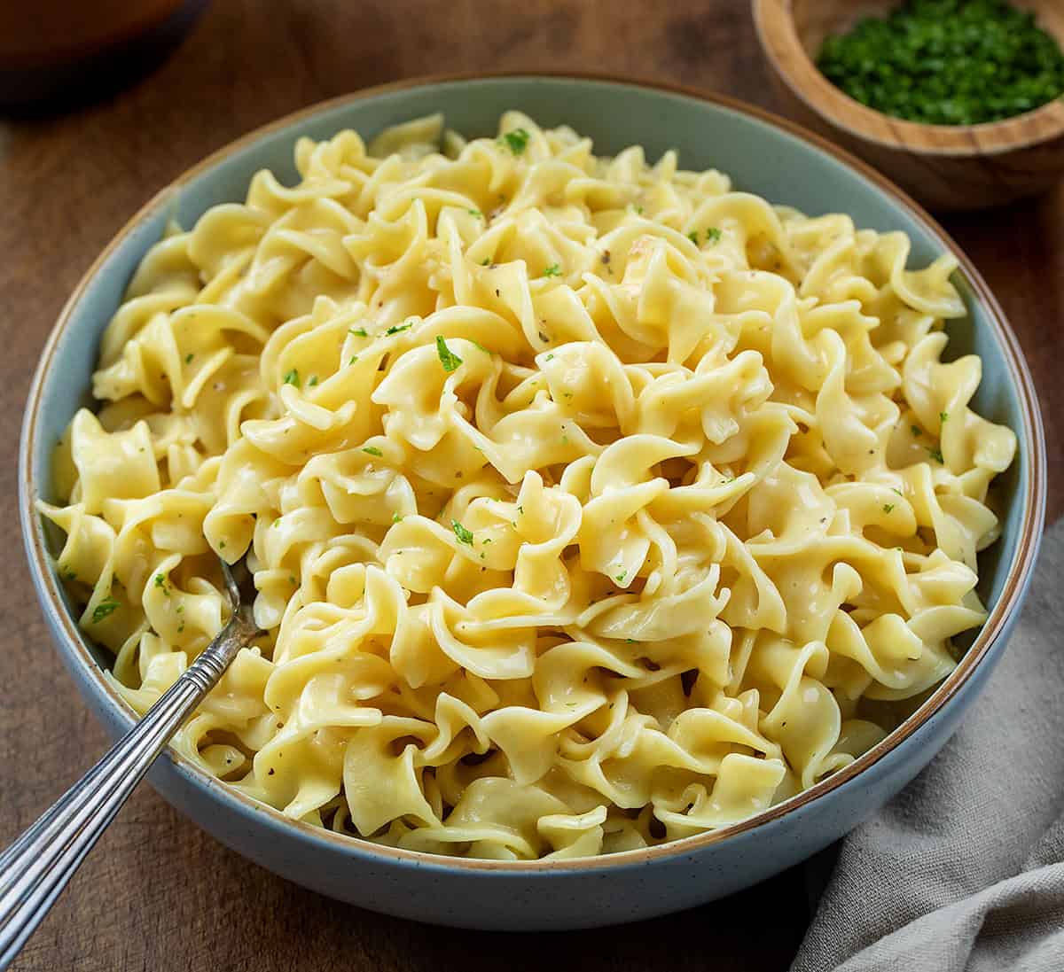 These Cafeteria Noodles are a simple, cozy one-pot recipe made with egg noodles simmered in chicken broth, then finished with cream of chicken soup, butter, and seasonings. They’re rich, creamy, and nostalgic, just like the comforting noodles you remember from school lunches. Close up of a blue bowl filled with Cafeteria Noodles and a spoon on a wooden table.