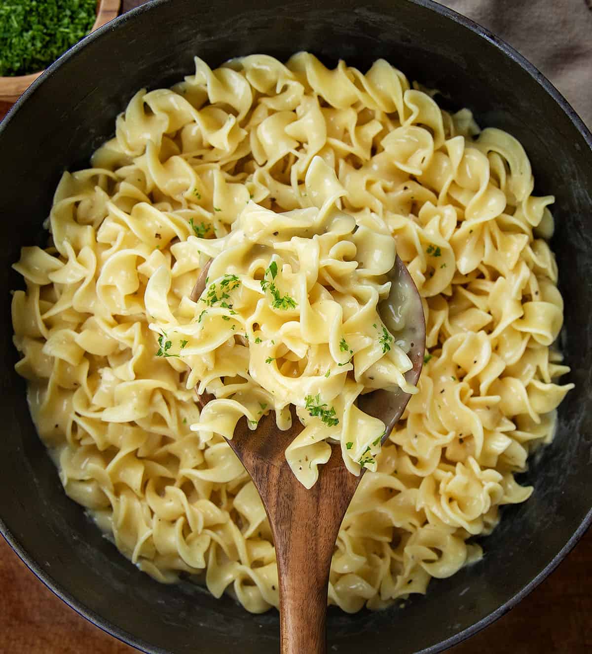 These Cafeteria Noodles are a simple, cozy one-pot recipe made with egg noodles simmered in chicken broth, then finished with cream of chicken soup, butter, and seasonings. They’re rich, creamy, and nostalgic, just like the comforting noodles you remember from school lunches. Pot of Cafeteria Noodles on a wooden table with a wooden spoon holding some noodles above the pot.