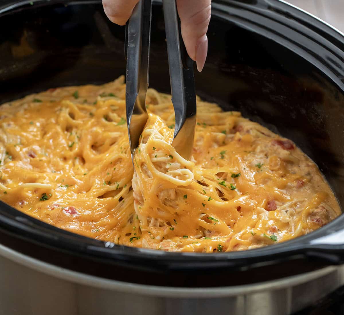 This Slow Cooker Cheesy Chicken Spaghetti is an easy, comforting dinner made with tender shredded chicken, a creamy cheese sauce, and perfectly cooked pasta! Tongs picking up a portion of Slow Cooker Cheesy Chicken Spaghetti from the crockpot.