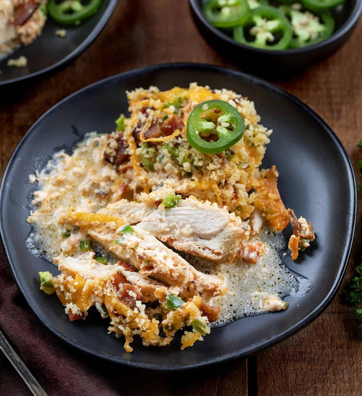 This Jalapeño Popper Chicken Bake is creamy, cheesy, and full of bold flavor in every bite. Tender chicken thighs are baked in a rich cream cheese sauce, then topped with crispy bacon, fresh jalapeños, and melty cheddar, all finished with a golden panko topping! Jalapeño Popper Chicken that has been partially cut up on a black plate.