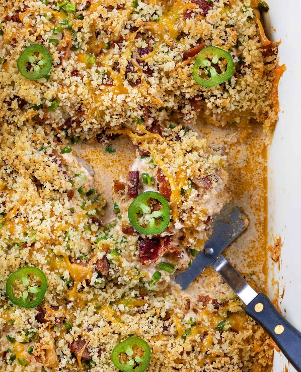 This Jalapeño Popper Chicken Bake is creamy, cheesy, and full of bold flavor in every bite. Tender chicken thighs are baked in a rich cream cheese sauce, then topped with crispy bacon, fresh jalapeños, and melty cheddar, all finished with a golden panko topping! Jalapeño Popper Chicken in a pan with a spatula.