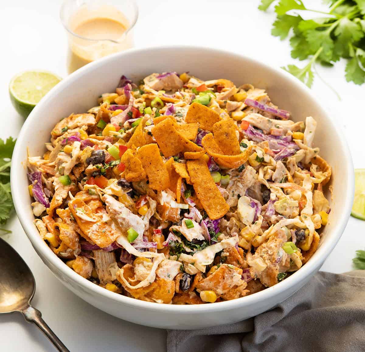 This Loaded Chicken Frito Cowboy Cabbage is a hearty, flavor-packed salad made with crunchy cabbage, shredded rotisserie chicken, black beans, corn, and fresh veggies, all tossed in a creamy chipotle dressing and finished with Chili Cheese Fritos! White bowl filled with Loaded Chicken Frito Cowboy Cabbage on a white table with fresh lime and cilantro.