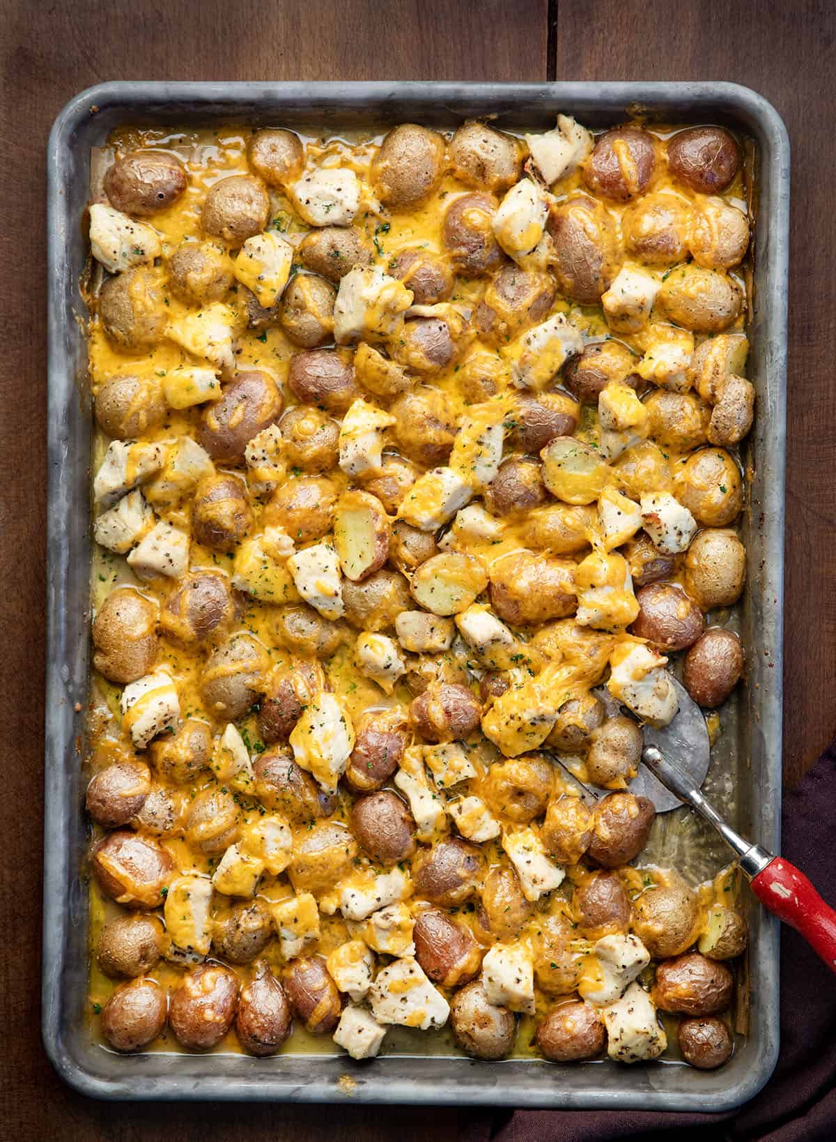 This One Pan Ranch Chicken and Potatoes is an easy sheet pan dinner made with tender chicken, roasted potatoes, and bold ranch seasoning, all finished with melted cheddar cheese! Sheetpan with Ranch Chicken and Potatoes on it and a spatula with a red handle.