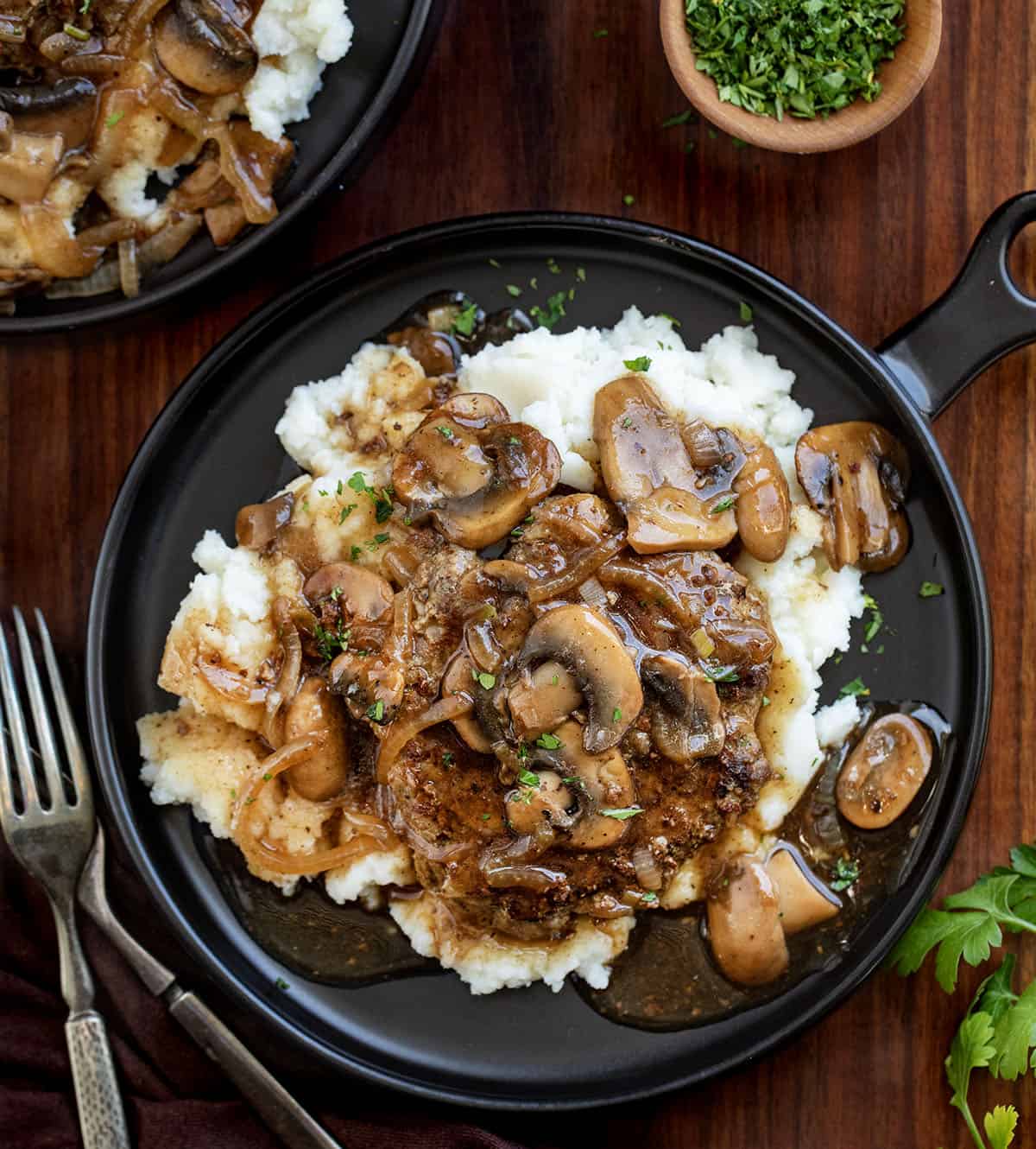 Poor Man’s Salisbury Steak with Mushrooms is an easy, budget-friendly dinner made with tender beef patties and a rich onion-mushroom gravy. It has all the classic comfort food flavor, with mushrooms adding even more depth to the savory sauce! Salisbury Steak with Mushrooms on plates with mashed potatoes on a wooden table from overhead.