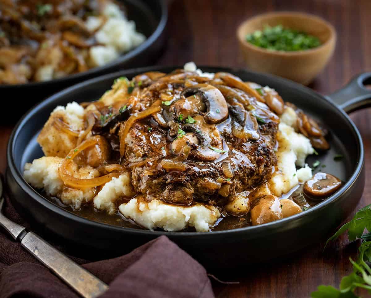 Close up of Salisbury Steak with Mushrooms on a bed of mashed potatoes on a black plate.
