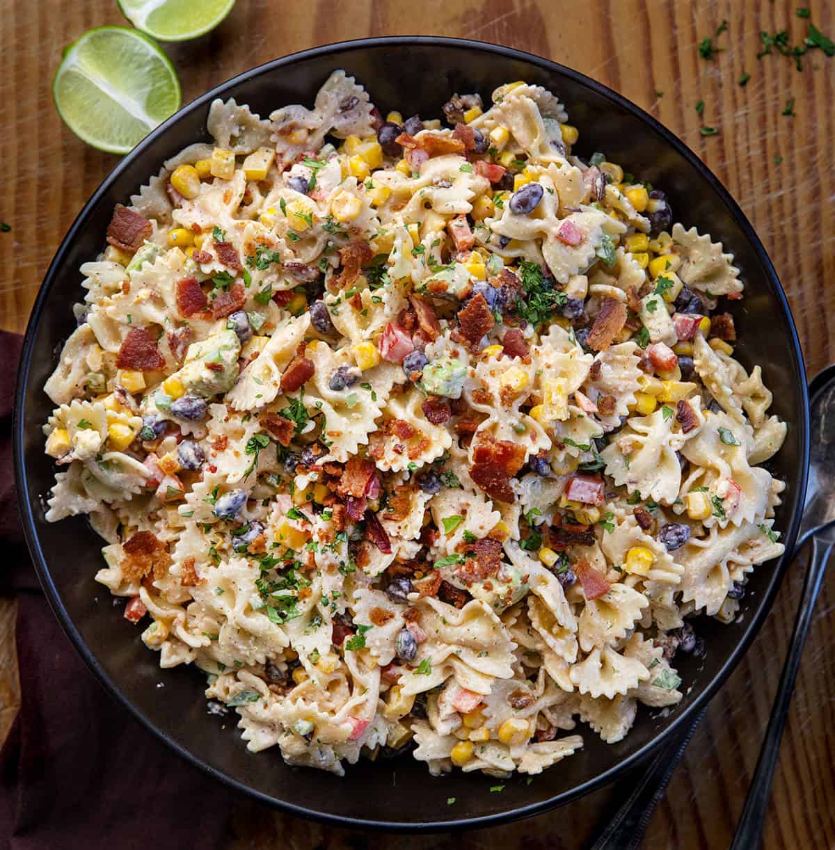This Southwest Pasta Salad is bold, creamy, and packed with flavor in every bite. Made with bowtie pasta, black beans, corn, bacon, fresh veggies, and a chili-lime dressing, it’s an easy make-ahead dish that’s fresh, hearty, and full of Southwest flavor! Black bowl filled with Southwest Pasta Salad on a wooden table from overhead.