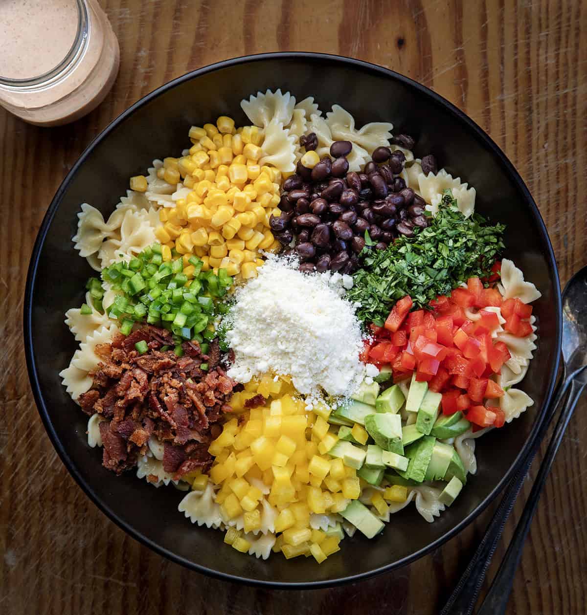This Southwest Pasta Salad is bold, creamy, and packed with flavor in every bite. Made with bowtie pasta, black beans, corn, bacon, fresh veggies, and a chili-lime dressing, it’s an easy make-ahead dish that’s fresh, hearty, and full of Southwest flavor! Raw ingredients for Southwest Pasta Salad in a black bowl on a wooden table before being mixed together.