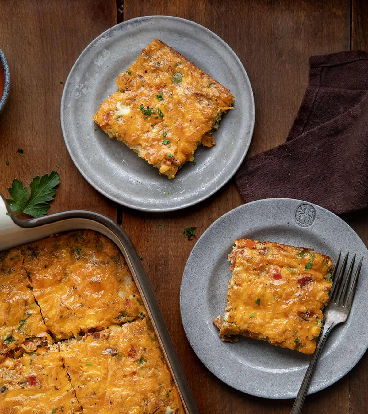 This Western Omelette Breakfast Bake is a quick, family-friendly breakfast that combines fluffy eggs, smoky bacon, sautéed peppers and onions, and melty cheddar cheese, all baked in one dish! Pieces of Western Omelette Breakfast Bake on plates next to the pan on a wooden table from overhead.