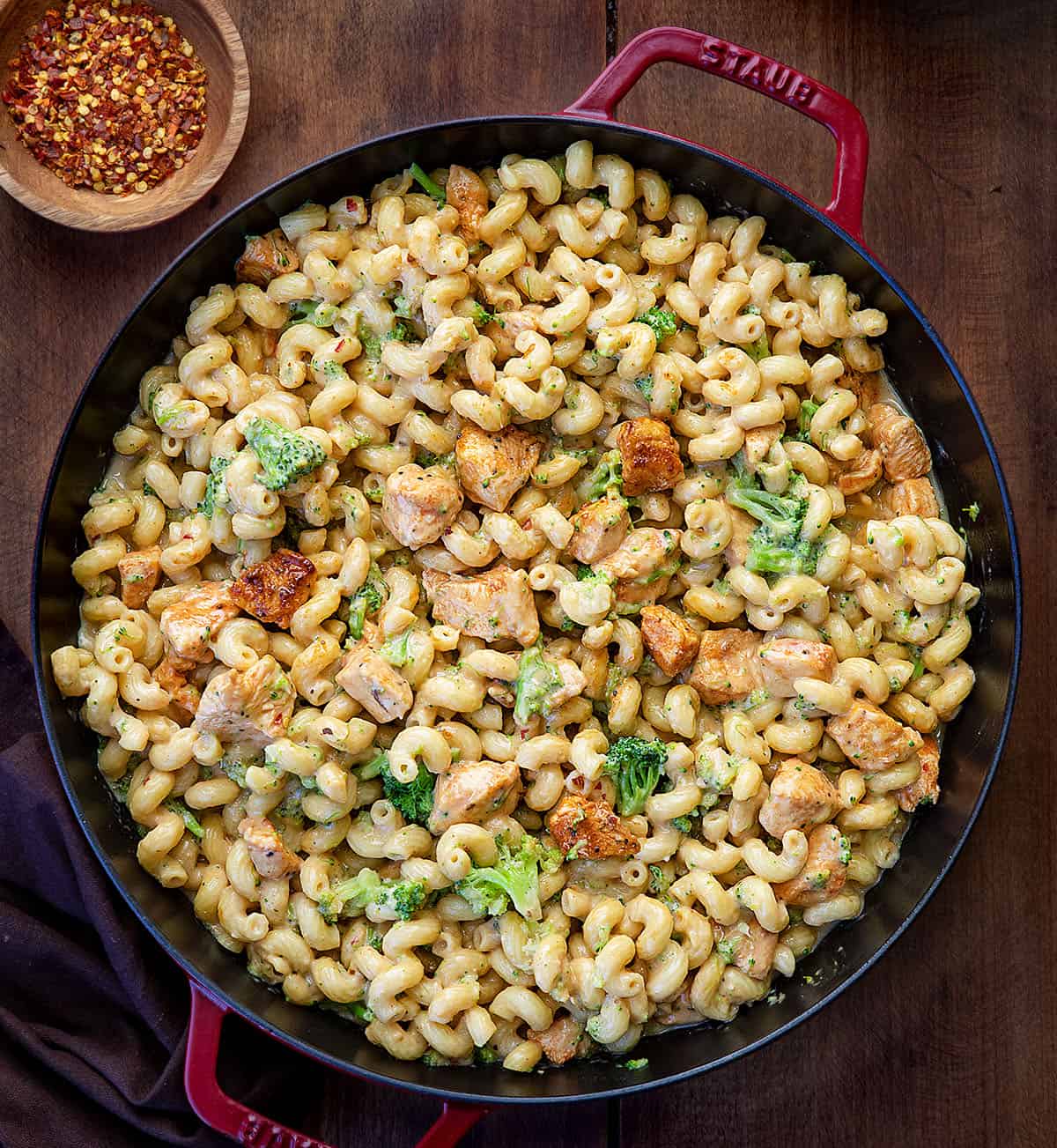 This White Cheddar Chicken Pasta with Broccoli is a creamy, comforting dinner made with tender chicken, cavatappi pasta, and crisp-tender broccoli all coated in a smooth white cheddar cheese sauce! Skillet of White Cheddar Chicken Pasta with Broccoli on a wooden table from overhead.