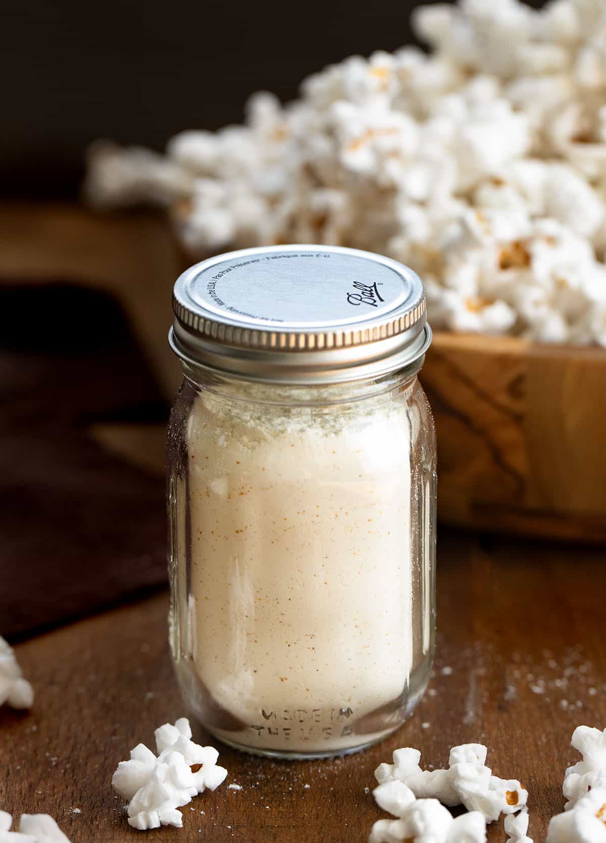 A quick and easy way to make your popcorn cheesy, flavorful, and irresistible. Just mix the spices and cheddar powder, shake on your favorite snack, and enjoy a homemade savory treat that’s ready in minutes! Jar of White Cheddar Popcorn Seasoning on a wooden table with popcorn in the background.