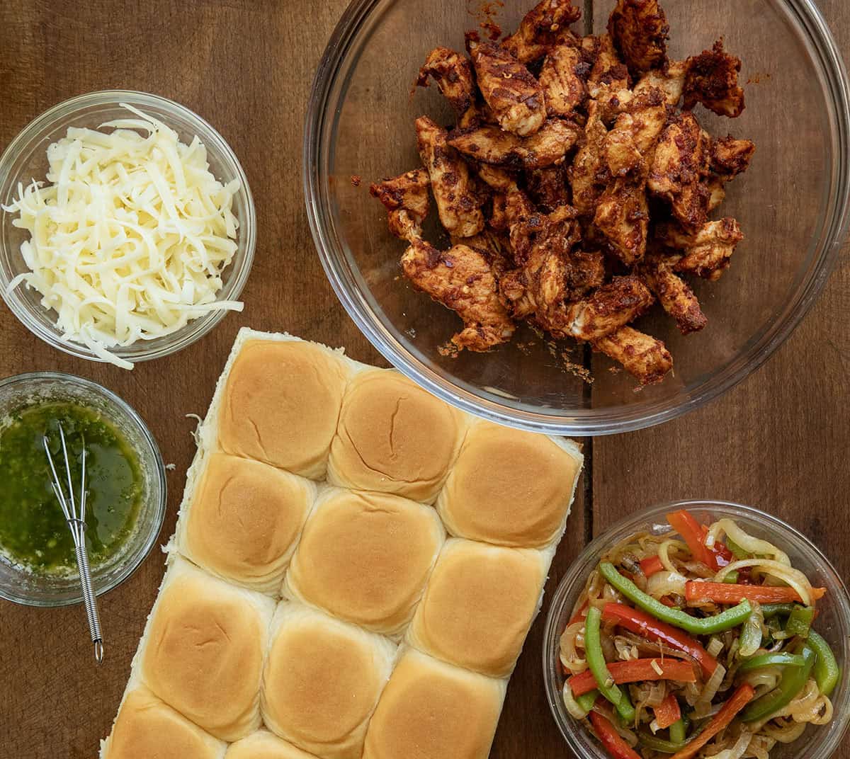 These chicken fajita sliders are loaded with juicy seasoned chicken, sautéed peppers and onions, melty cheese, and buttery Hawaiian rolls baked until golden brown. Ingredients to make Chicken Fajita Sliders on a wooden table from overhead.