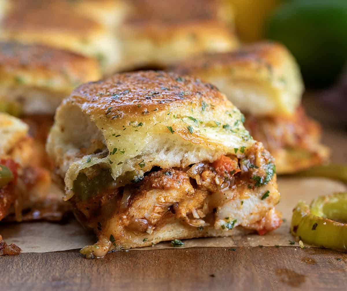 These chicken fajita sliders are loaded with juicy seasoned chicken, sautéed peppers and onions, melty cheese, and buttery Hawaiian rolls baked until golden brown. Close up of Chicken Fajita Slider on a wooden table with more sliders in the background.