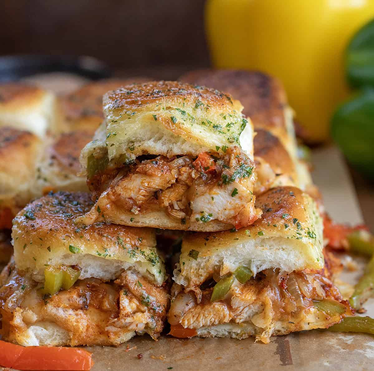 These chicken fajita sliders are loaded with juicy seasoned chicken, sautéed peppers and onions, melty cheese, and buttery Hawaiian rolls baked until golden brown. Chicken Fajita Sliders stacked on a wooden table with bell peppers in the background.