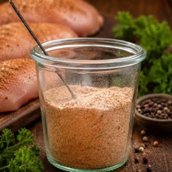 Jar of Chicken Seasoning on a wooden table with raw chicken and peppercorns and a measuring spoon in the jar.