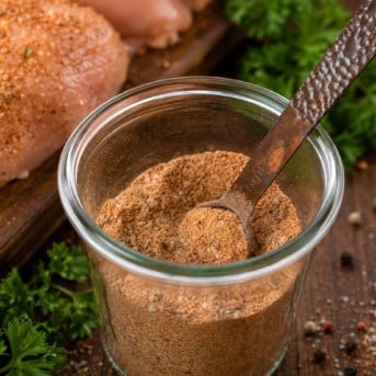 Jar of Chicken Seasoning on a wooden table with raw chicken and peppercorns and a measuring spoon in the jar.