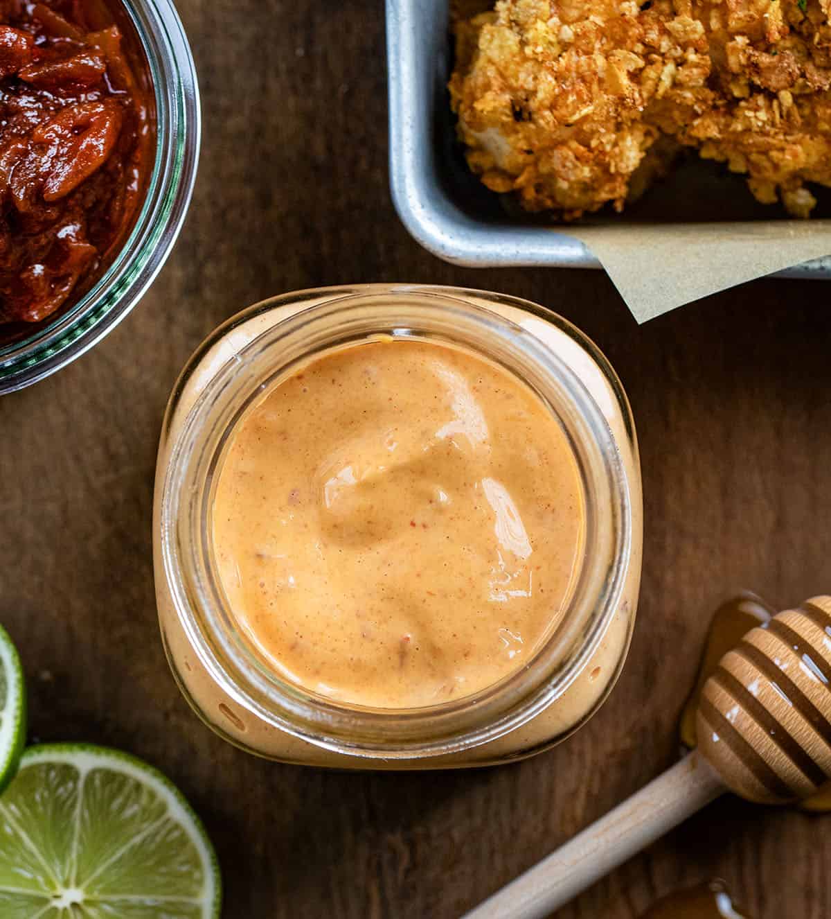 This Homemade Chipotle Mayo is creamy, smoky, and just the right amount of spicy with a touch of tangy lime and subtle sweetness. Looking down on a jar of Chipotle Mayo on a wooden table surrounded by honey, lime, chipotle peppers, and chicken.