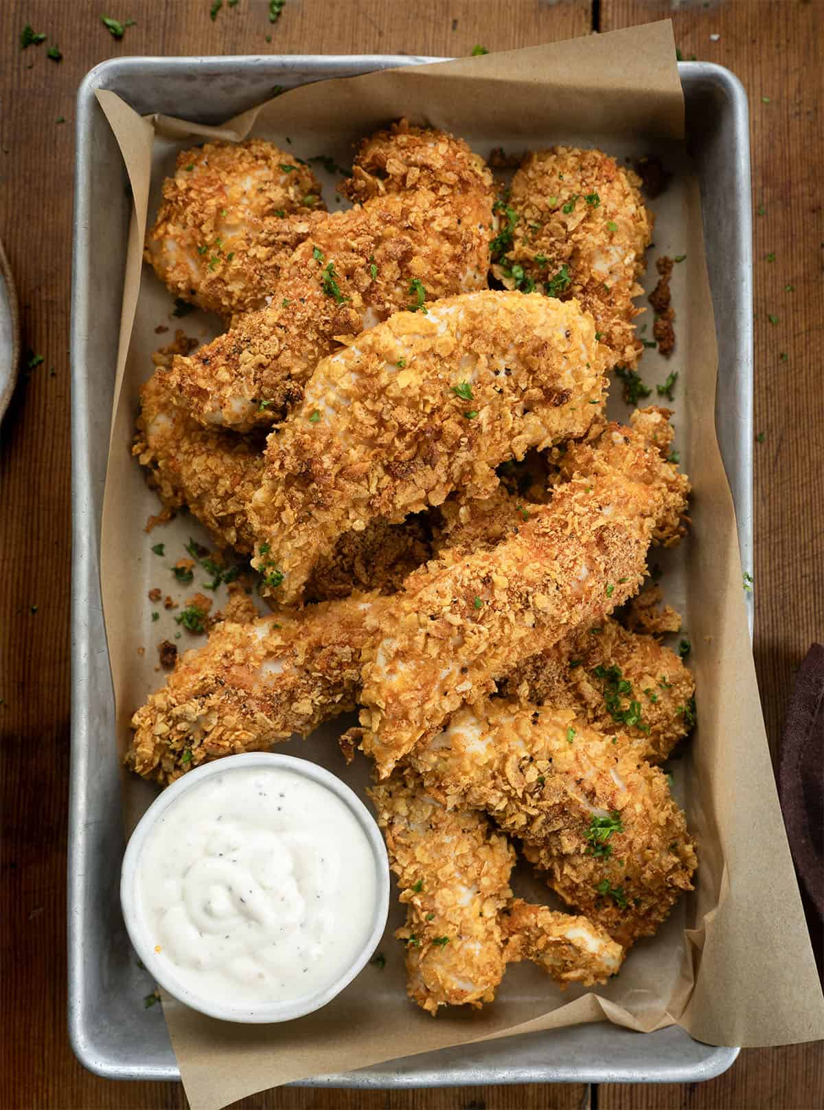 This Cornflake Chicken is crispy, golden, and baked, not fried, making it an easy weeknight dinner the whole family will love. Simple pantry ingredients come together for a no-fuss meal that’s big on flavor and short on prep time! Pan of Cornflake Chicken with some ranch dressing on a wooden table.