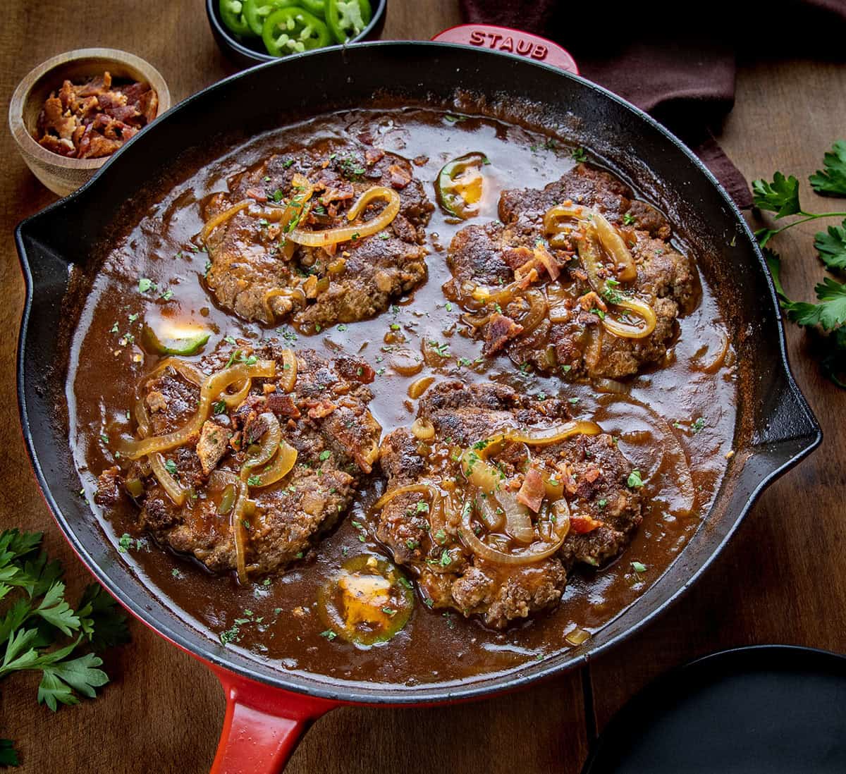 This Cowboy Salisbury Steak is a hearty, flavor-packed dinner made with juicy beef patties and a rich onion gravy with BBQ sauce, bacon, and a little jalapeño heat. Red skillet of Cowboy Salisbury Steak on a wooden table.