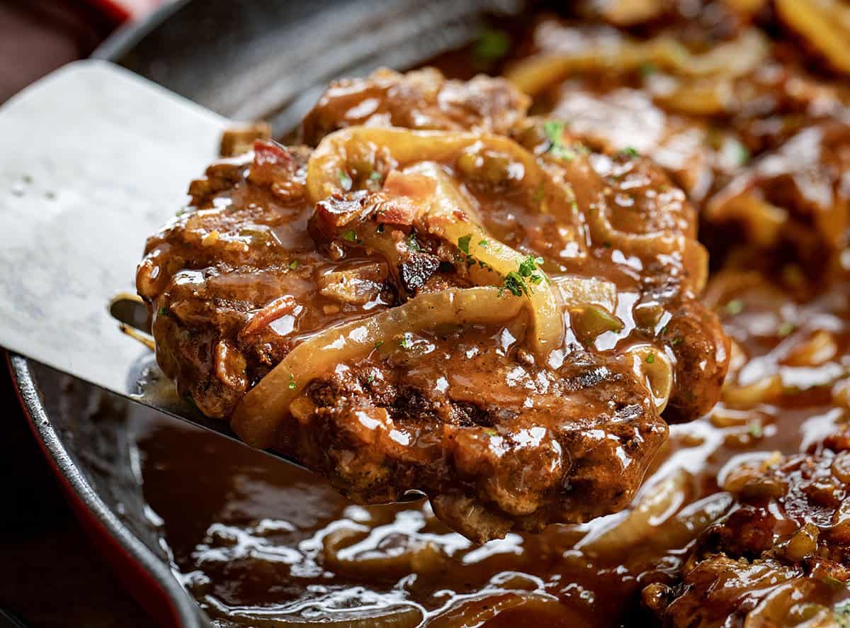 This Cowboy Salisbury Steak is a hearty, flavor-packed dinner made with juicy beef patties and a rich onion gravy with BBQ sauce, bacon, and a little jalapeño heat. Spatula picking up a piece of Cowboy Salisbury Steak from the pan.