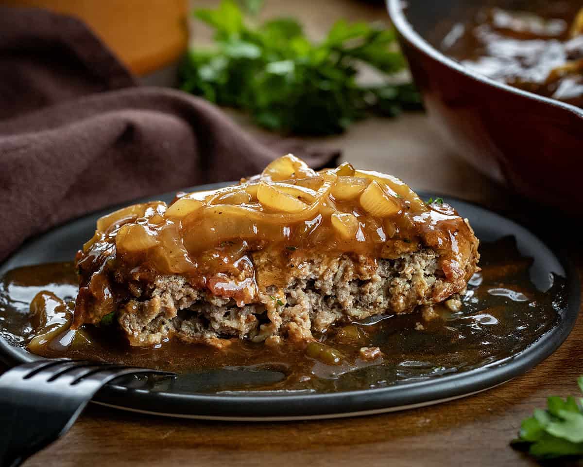 This Cowboy Salisbury Steak is a hearty, flavor-packed dinner made with juicy beef patties and a rich onion gravy with BBQ sauce, bacon, and a little jalapeño heat. One piece of Cowboy Salisbury Steak with half eaten on a black plate.