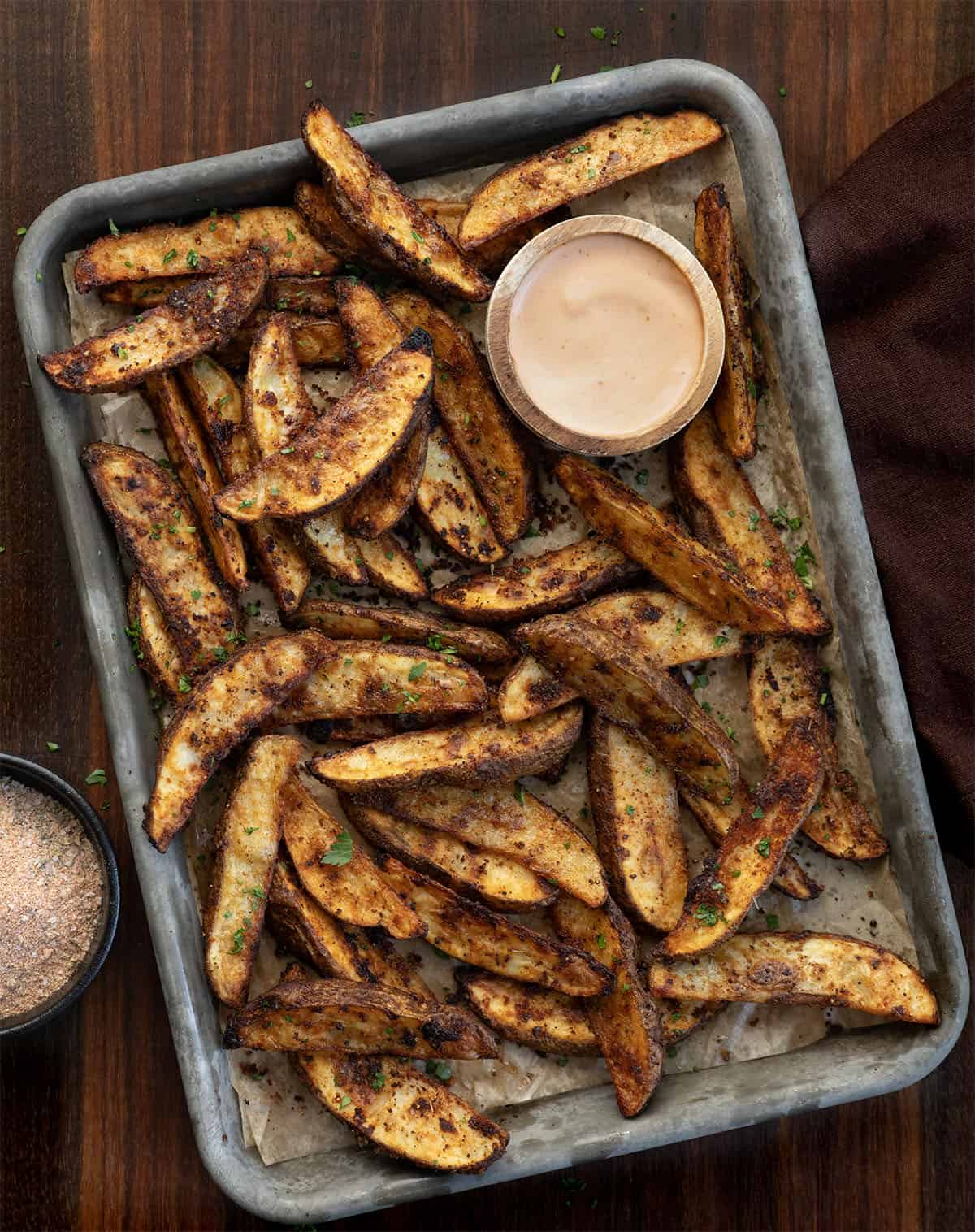 Sheet pan with Crispy Potato Wedges and dipping sauce on it on a wooden table from overhead.